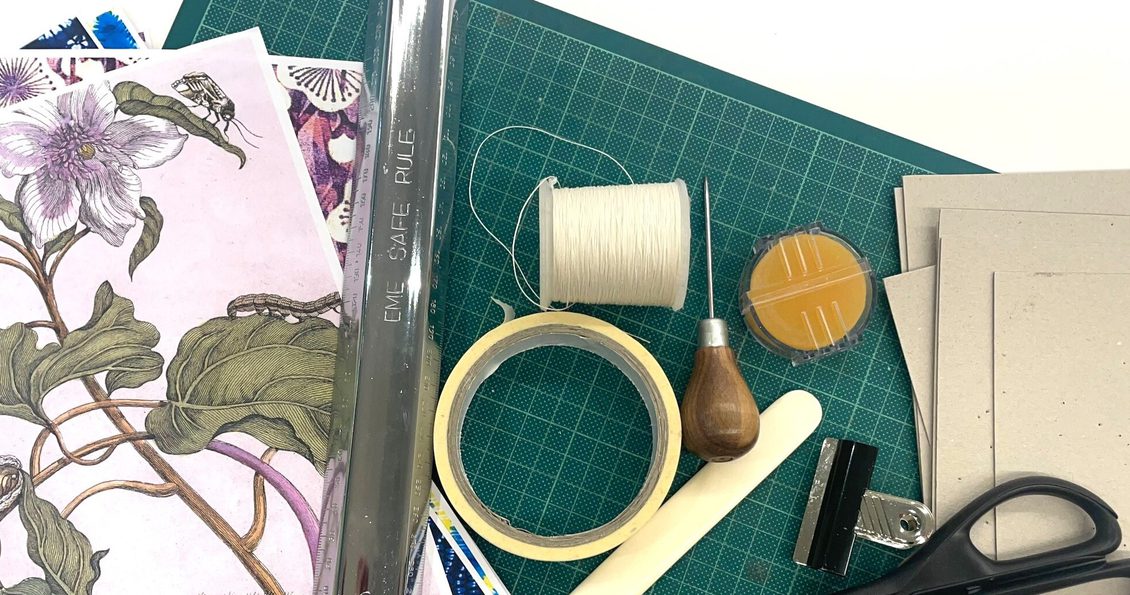 Bookmaking tools spread on a green mat. The tools include pink floral paper, a metal ruler, a roll of masking tape, a spool of white thread, an awl, an orange sponge, a white paper creaser, brown paper, a bulldog clip and scissors.