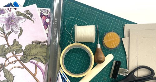 Bookmaking tools spread on a green mat. The tools include pink floral paper, a metal ruler, a roll of masking tape, a spool of white thread, an awl, an orange sponge, a white paper creaser, brown paper, a bulldog clip and scissors.