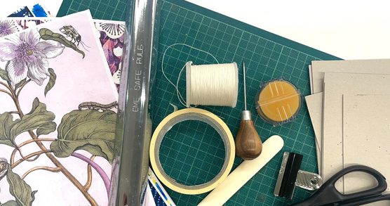 Bookmaking tools spread on a green mat. The tools include pink floral paper, a metal ruler, a roll of masking tape, a spool of white thread, an awl, an orange sponge, a white paper creaser, brown paper, a bulldog clip and scissors.