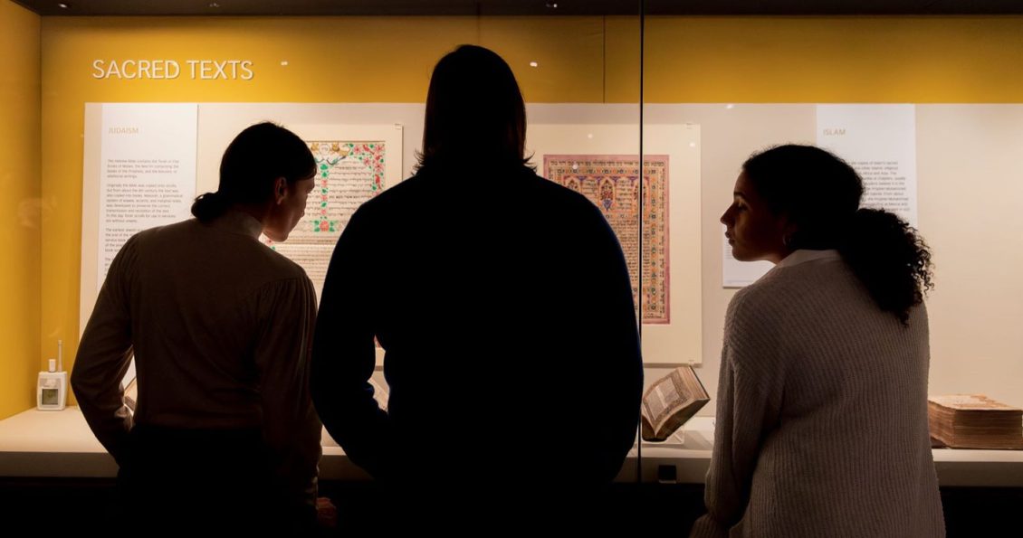 Three people are silhouetted against a brightly lit display case as they look at the books instead. The case has a yellow background.