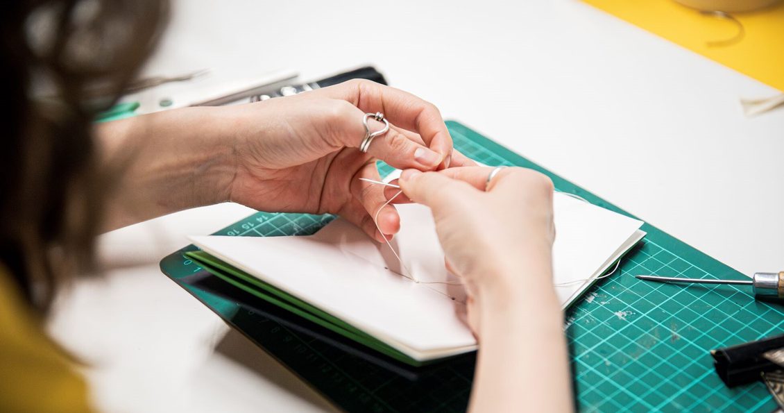 Hands sewing thread through paper to make a notebook.