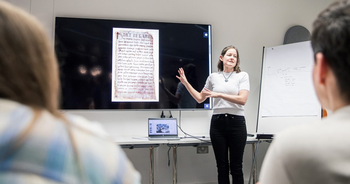 A woman stood in front of a screen displaying an Old English manuscript talks to two people sat in a classroom-style environment.