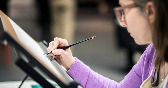 A woman wearing glasses and a purple jumper using a calligraphy pen to write on a drawing board.