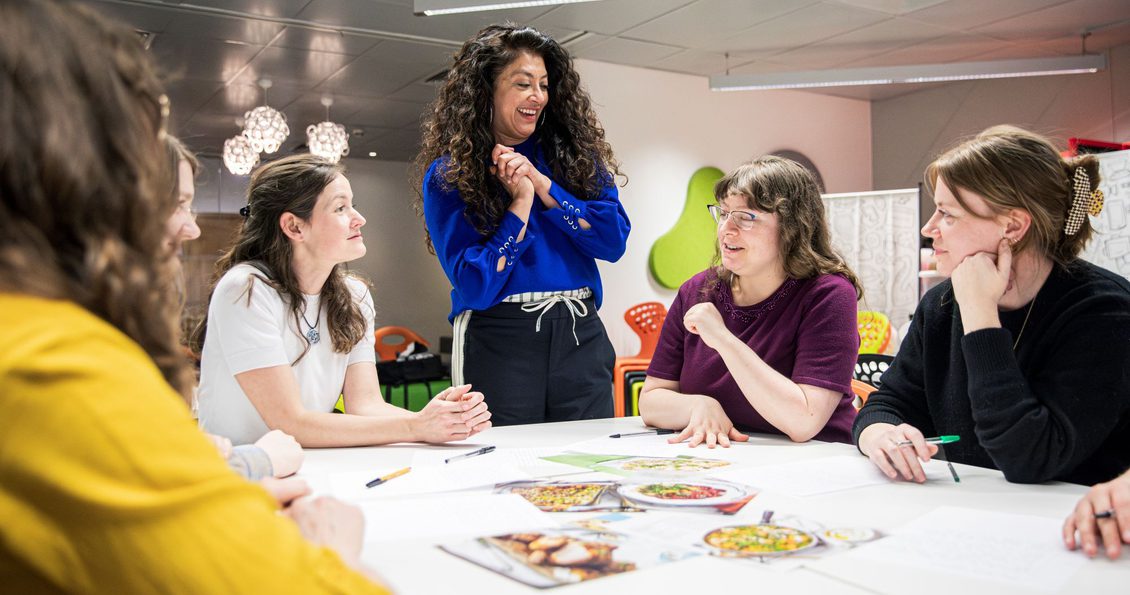 Image shows course tutor, Mallika Basu, standing at a table with four course participants, there are images from food magazines on the table, the group are discussing food photography