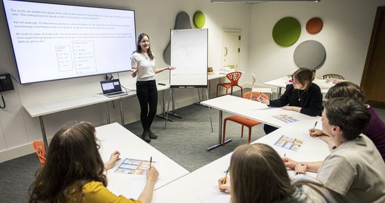 Image shows a group of adult learners seated at tables looking at a screen and listening to an educator