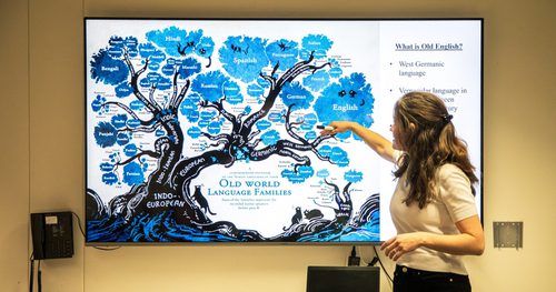 A woman gesturing to a large screen behind her which shows an illustration of a blue tree and outlines elements of the Old English language.