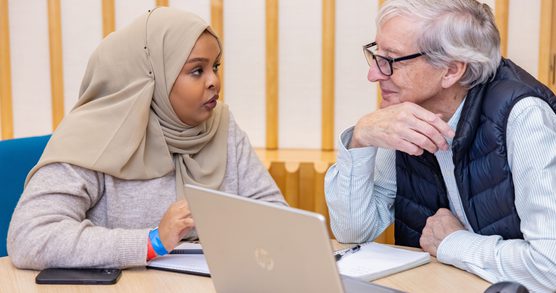 Two people sat next to each other talking in front of a laptop.
