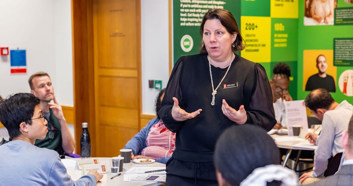 Woman stood up delivering a workshop, with people sat around tables listening.