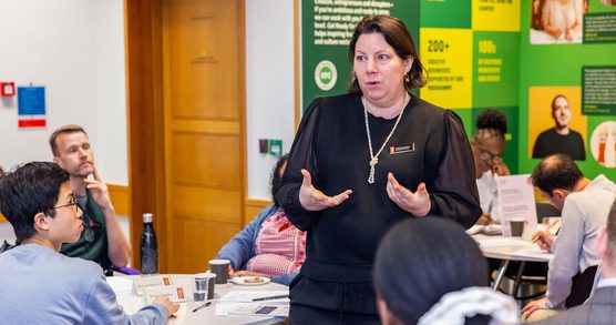 Woman stood up delivering a workshop, with people sat around tables listening.