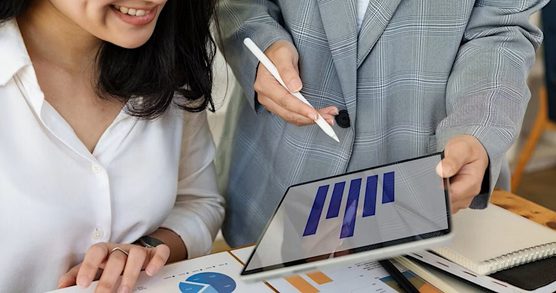 A view of a tablet being held by a person in a grey suit jacket in order that someone with dark hair in a white shirt can view the screen