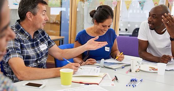 A group of adults say around a table. They are talking animatedly with each other. There are pens and papers on the table.