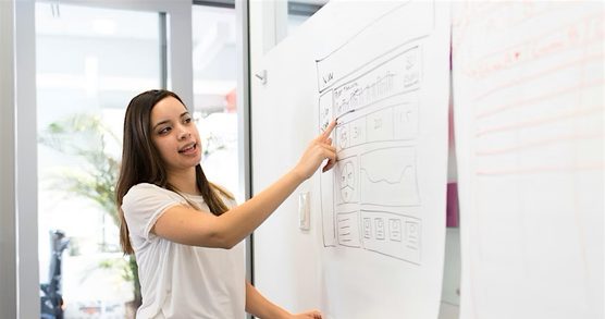 Woman with long brown hair, wearing a white T-shirt, points at a sheet of paper pinned to a wall. The paper is covered in diagrams