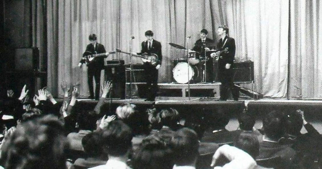 Black and white photo of the The Beatles performing on a stage at Stowe School, students form a crowd in front of the stage.