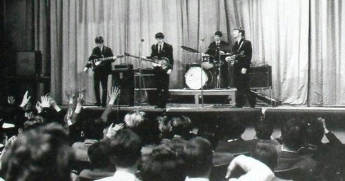 Black and white photo of the The Beatles performing on a stage at Stowe School, students form a crowd in front of the stage.
