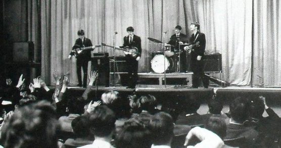 Black and white photo of the The Beatles performing on a stage at Stowe School, students form a crowd in front of the stage.