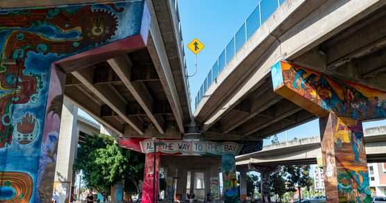 The image shows an urban scene beneath a large highway overpass on a clear, sunny day. Multiple concrete freeway ramps intersect overhead, creating a layered structure of beams and pillars.