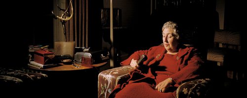 Agatha Christie in old age, wearing a red dress, sat in an armchair. Her eyes are closed. Beside her is a table packed with an assortment of objects.