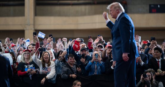 President Donald Trump, a white eighty-year-old man in a blue suit, walks past a crowd barrier at an indoor political rally while supporters behind the barrier wave, cheer, and hold up smartphones to take photos. Several people wear red caps and patriotic clothing. Photographers kneel near the front of the crowd, and the background shows the interior of a large event venue. The public figure is slightly out of focus in the foreground, while the crowd is sharply focused.