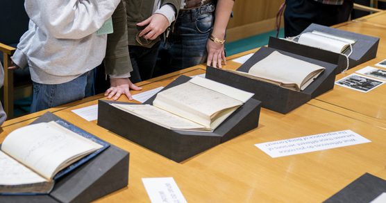Four open books on display stands sit on a wooden table. Four people, whose upper bodies are out of shot, stand next to the table.