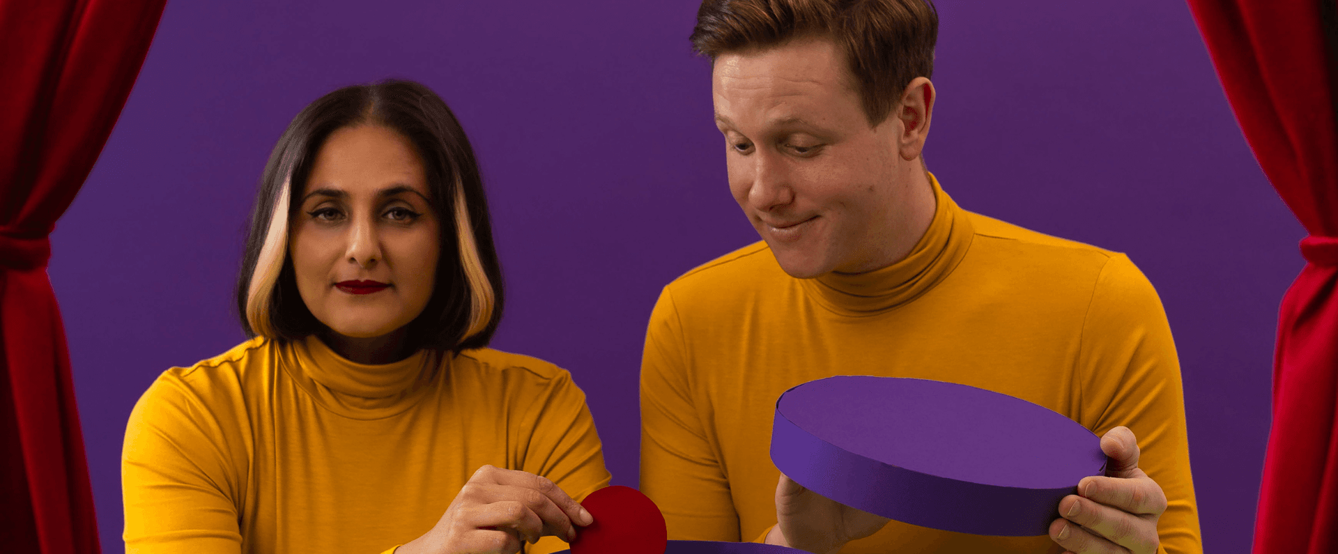 Two people dressed in yellow; one is holding the lid of a cut-out purple biscuit tin, the other is holding a cut-out red circular biscuit. They are against a purple background with red curtains to either side.