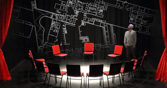 A circle of red chairs, and Seth Kriebel standing, in front of a large architectural drawing of the British Library.