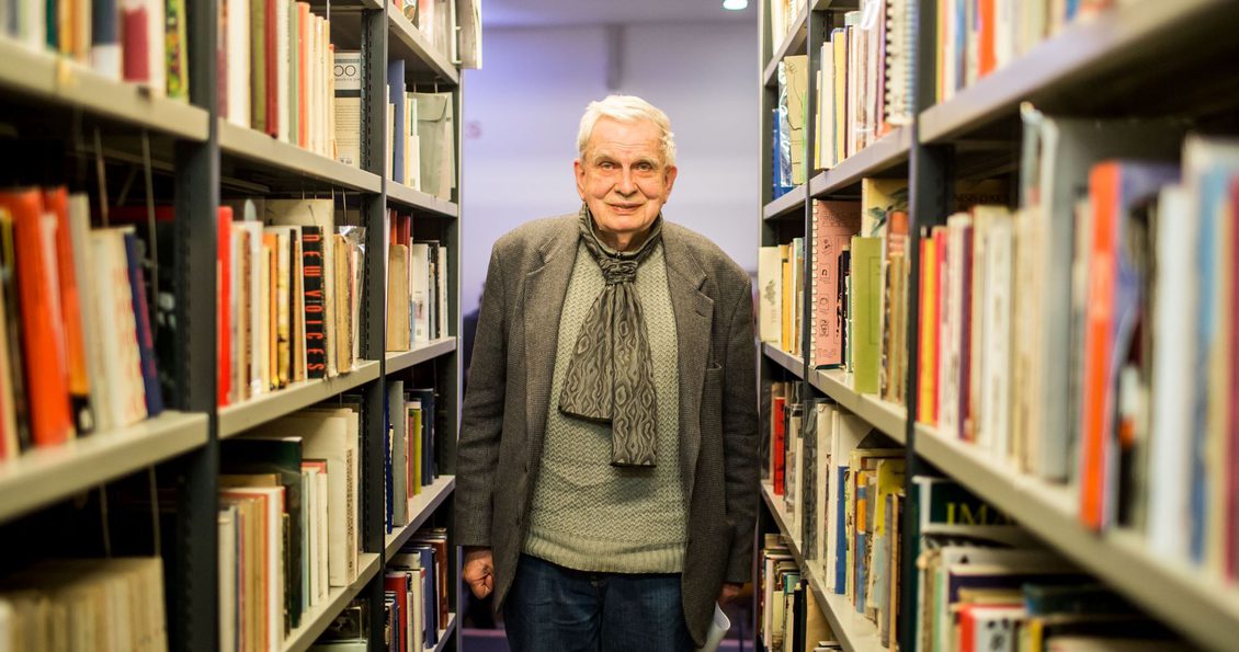 Photo of Tomas Venclova in a jumper, scarf and jacket. He is standing between two shelves of Library books.