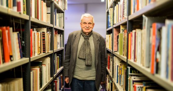 Photo of Tomas Venclova in a jumper, scarf and jacket. He is standing between two shelves of Library books.
