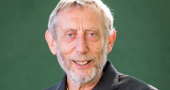 A smiling man with grey hair, beard and moustache, wearing a suit jacket with a blue jumper and stripy t-shirt underneath