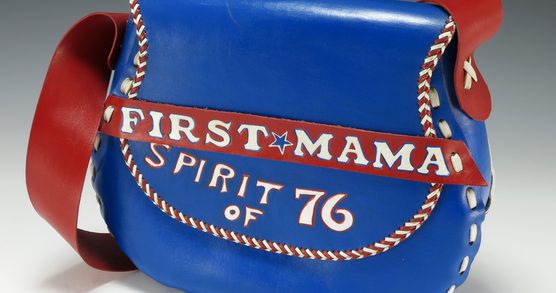Blue leather shoulder bag with red straps and white lacing, styled like a baseball glove, featuring the words “FIRST MAMA” and “SPIRIT OF ’76” in red and white lettering across the front.