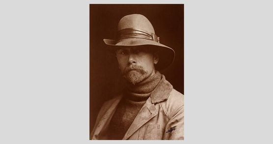 Head and shoulders sepia toned photo of a bearded man wearing a polo neck sweater, a blazer with a cord collar, and a hat.