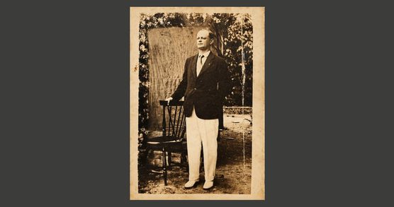 Outdoors photograph of a man wearing a dark jacket, shirt and tie and white trousers holds onto the back of a wooden chair.