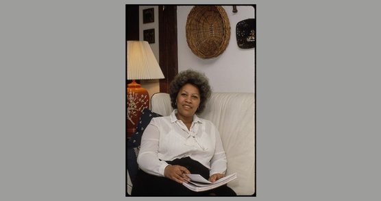 Toni Morrison sits on a white couch, wearing a white long-sleeve blouse and dark skirt, holding papers and a pen. Behind her are a large lamp and works of art on the wall.