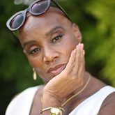 Photograph of Andi Oliver, head in hand, sunglasses on head, white top, gold jewellery