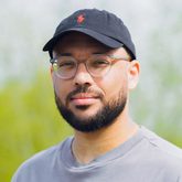 A bearded man wearing glasses, a grey cap and light grey t-shirt.