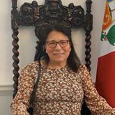 Photo of Constantina Higbie, patterned top, seated on a wooden chair, shoulder length dark hair, glasses