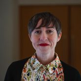 Image shows a profile image of Elena Medel. Elena is a white woman with short brown hair. She is wearing a white, orange, green and black patterned collared shirt and a black cardigan.