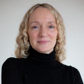 A woman with blonde curly hair, wearing a black polo neck top with her arms folded