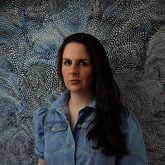 Photo of Karina Lickorish Quinn. Long dark hair, blue denim dress, gold hoop earrings, blue patterned background.