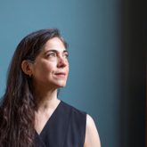 Photo of Marjorie Lotfi, standing against a blue background, long dark hair across one shoulder, black top, gold earrings