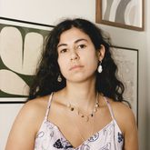 Photo of Monika Radojevic, long wavy dark hair, dangly earring, two necklaces, strappy pink top, artwork background