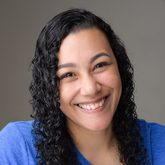 A smiling woman with long, curly hair wearing a bright blue t-shirt