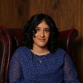 A woman with dark curly hair wearing a blue blouse, sitting in a red leather arm chair
