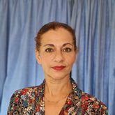 photo of Shara Atashi, looking confidently at camera, floral top, red lipstick, hair tied back, blue background