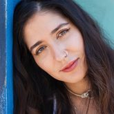 Photo of Nikita Gill, leaning against a blue wall, long dark hair