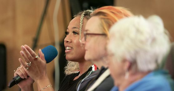 Three women sat in a row, all in profile, one is speaking into a microphone