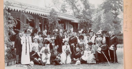 A black and white Edwardian era photograph showing a group of men and women posed in front of a vine-covered porch in India.