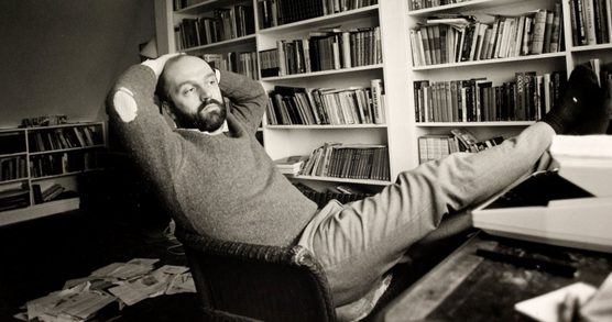 A bearded man sits back in a chair with his feet up on a desk, and arms lifted to his head. Behind him are full bookshelves.
