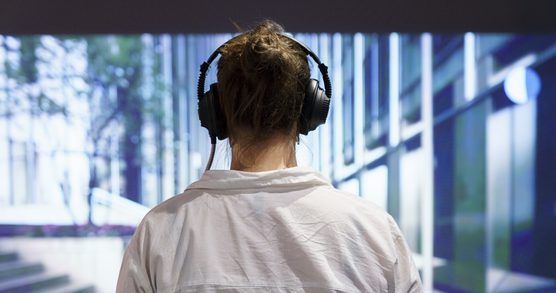 A woman with dark hair in a bun wearing a white shirt and over ear headphones stands in front of a large digital projection showing a building.