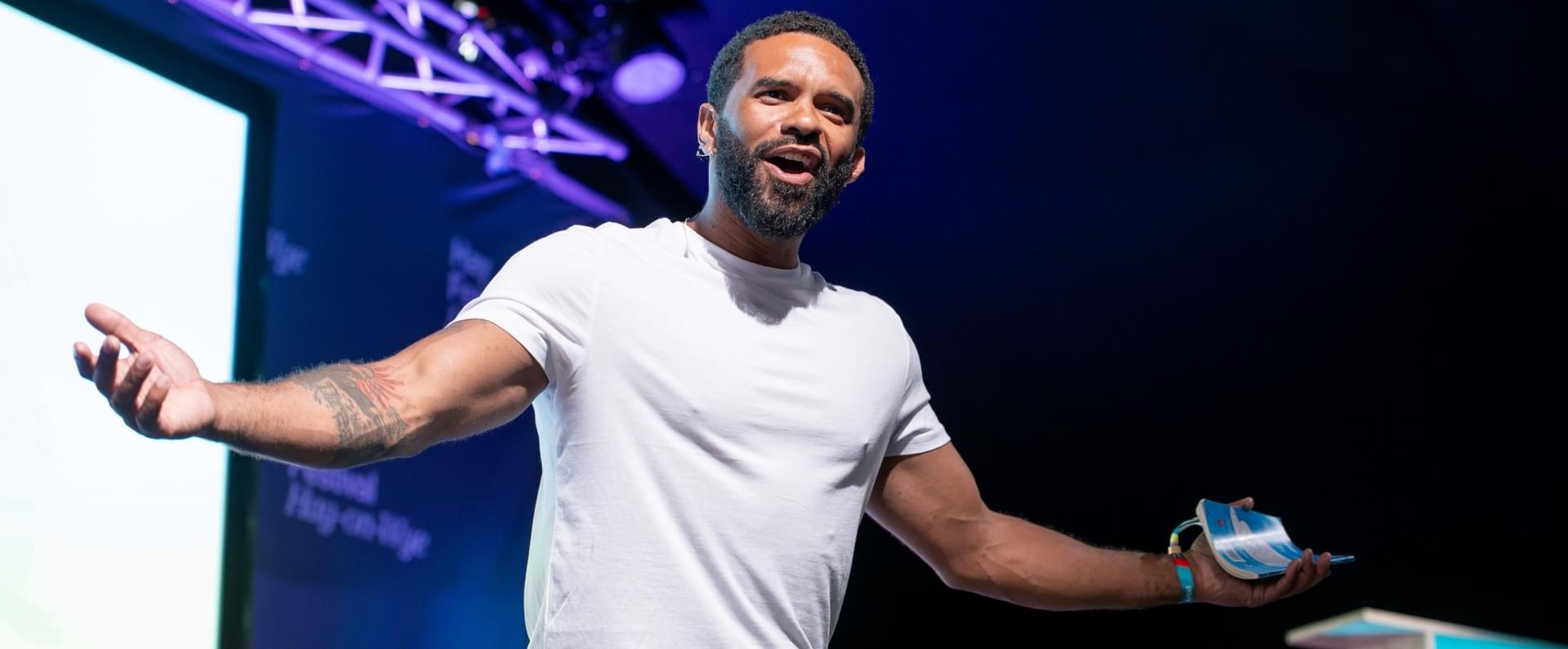 A man in a white t-shirt stands on a stage, talking enthusiastically to a crowd with his arms outstretched, holding a small book in one hand.