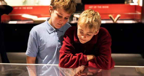 Two school boys, one in a blue polo shirt, the other in a red jumper, lean over an exhibition display case and to look inside of it. Their expressions show interest in what they are looking at.